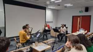 A group of four students playing on Adolphe Sax instruments in a lecture hall. They are sitting in quartet formation and playing as an SATB quartet. 