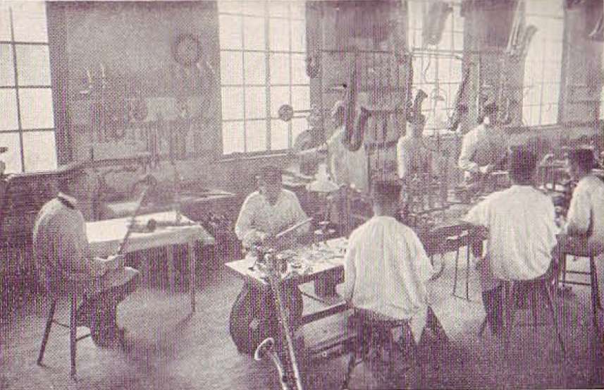 A faded photograph of several workers at a table in a factory hall, busy assembling a series of saxophones.